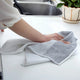A person's hand and arm are shown cleaning a white kitchen countertop with a gray and white textured microfiber cloth. In the background, a stainless steel sink and a potted green plant are visible.