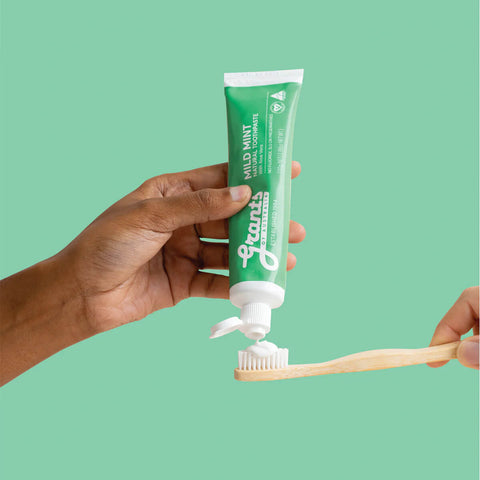 Hand squeezing toothpaste onto a bamboo toothbrush against a green background.