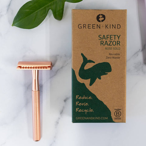 Rose gold safety razor next to its cardboard box on a marble surface with a leaf in the background.