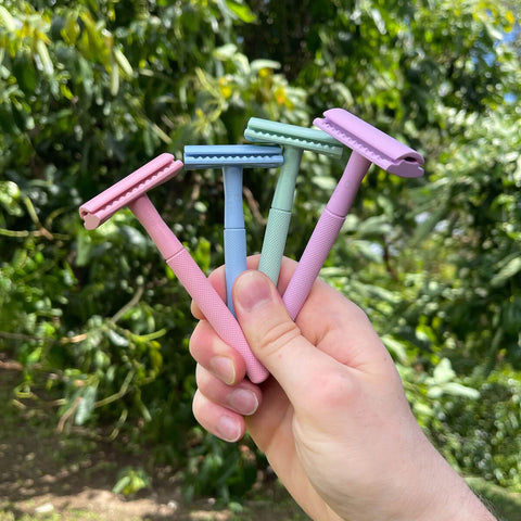 Hand holding four colorful razors against a green outdoor background.