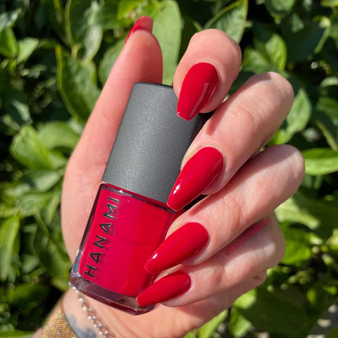 Hand holding a red nail polish bottle with green foliage in the background