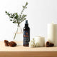 Bottle of hair styling product on a wooden surface with decorative elements like candles and plants.