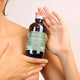 Honest Skincare Kawakawa Infused Sweet Almond Oil 250ml lifestyle shot of two hands holding the dark bottle beside a person's bare shoulder