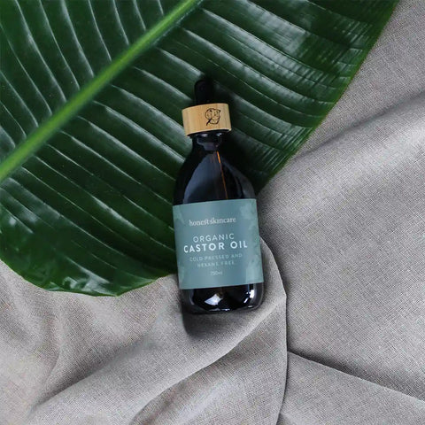 Honest Skincare Organic Castor Oil 250ml lifestyle shot of the dark bottle on a large green leaf over grey fabric, styled lifestyle photo