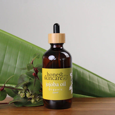 Honest Skincare Organic Jojoba Oil 250ml lifestyle shot of the dropper bottle on wood beside green leaves, styled lifestyle photo