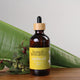 Honest Skincare Organic Jojoba Oil 250ml lifestyle shot of the dropper bottle on wood beside green leaves, styled lifestyle photo