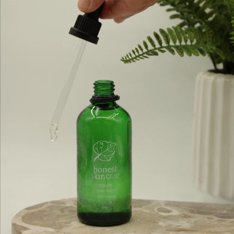 Honest Skincare Organic Rosemary Revitalising Hair Serum 100ml lifestyle shot of the green bottle on a stone base with a dropper lifted beside a plant