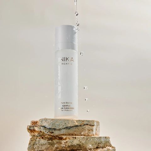 INIKA Organic Pure Biome Gentle Milk Cleanser 100ml lifestyle shot of the bottle on a stack of stones with water drops falling