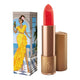 Lipstick next to box packaging that pictures a woman in a yellow frilly dress with a parrot in hand and sea views from a balcony. Gold lipstick case with flower detail on lid. Natural, clean cosmetics.