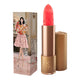 Lipstick next to box packaging that pictures a woman in a tiered pink dress holding a bouquet of flowers. Gold lipstick case with flower detail on lid. 