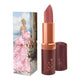 Lipstick next to box packaging that pictures a woman in a pale pink ruffle dress. Gold & red lipstick case with flower detail on lid. 