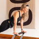 Woman in black athletic wear performing a yoga pose on a wooden floor with a decorative tapestry in the background.