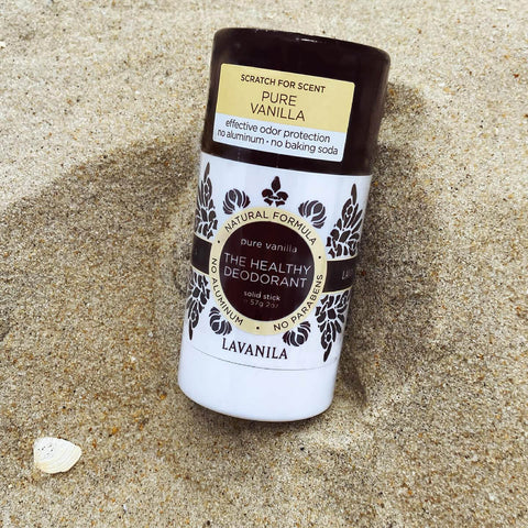 Lavanila The Healthy Deodorant Pure Vanilla lifestyle shot of the stick standing in beach sand beside a small shell, close up