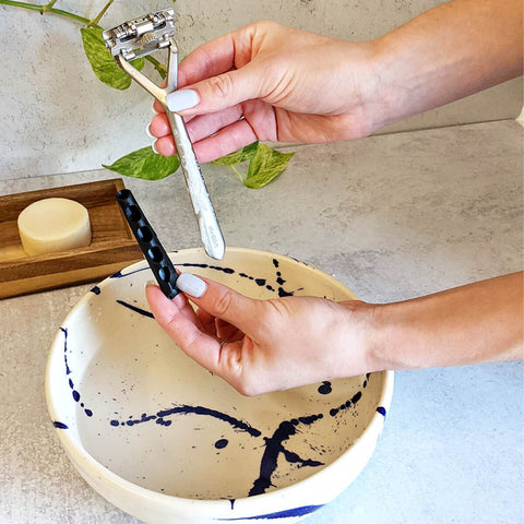 Person holding a razor over a bowl with a soap bar and leaves on a light surface