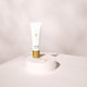 Lily Lolo Hydrating Face Primer 25ml lifestyle shot of the white and gold tube on a white round plinth against a pale backdrop