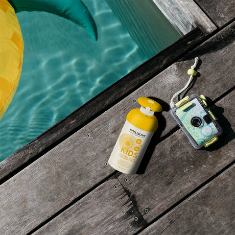 A bottle of sunscreen and a waterproof camera resting on a wooden deck next to a swimming pool.