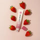 A tube of Little Urchin Strawberry Lip Balm is arranged on a light pink background, surrounded by fresh strawberries.