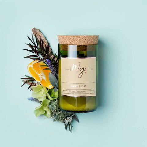 Mojo Candle Co. Wine Bottle Soy Wax Candle Oakmoss & Sage lifestyle shot of the bottle candle among citrus slices and leaves on an aqua backdrop