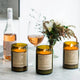 Mojo Candle Co. Wine Bottle Soy Wax Candle Oakmoss & Sage lifestyle shot of three lit bottle candles beside glasses of rosé and a vase of flowers