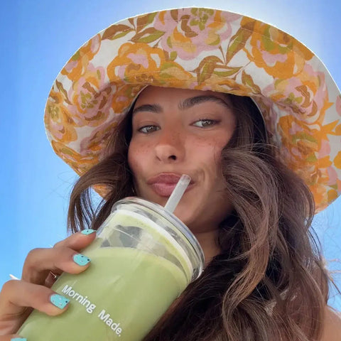 Morning Made Matcha Latte Mango Blend 300g lifestyle shot of a woman in a printed bucket hat sipping a green drink through a straw