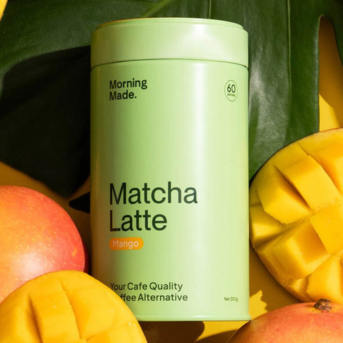 Morning Made Matcha Latte Mango Blend 300g lifestyle shot of the green tin among ripe mangoes and large green leaves, styled lifestyle photo