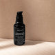 A black pump bottle of MUKTI Golden Glow Bronzing Drops placed on a light wood surface against a textured beige background.