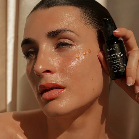 A close-up of a woman holding a black bottle of MUKT Gold Glow Golden Enhancing Drops next to her cheek where three golden drops of serum are applied, showing a glowing, hydrated complexion.