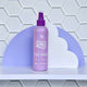Purple 200ml spray bottle of No Nasties Slick Kids Alcohol Free Hair Spray, against a white cloud structure and purple background elements. 