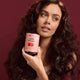 A woman with long, wavy dark brown hair holds a pink tin of Hair Ritual hair oil against a solid dark burgundy background.