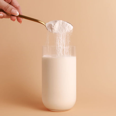 Spoon pouring Nutra Organics Organic Whey Protein - Vanilla 750g powder into a glass of milk on a beige background.