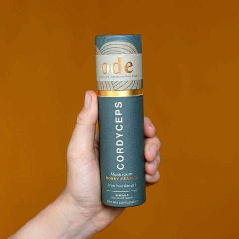 Hand holding tube of Cordyceps Mushroom Honey Pearls against an orange background. 