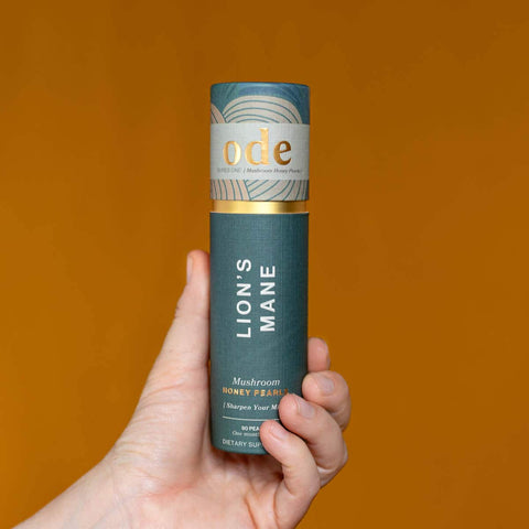 Hand holding tube of Lion's Mane Mushroom Honey Pearls against an orange background. 
