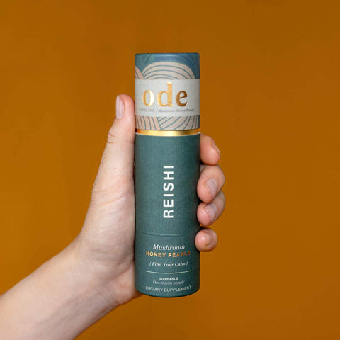 Hand holding tube of Reishi Mushroom Honey Pearls against an orange background. 
