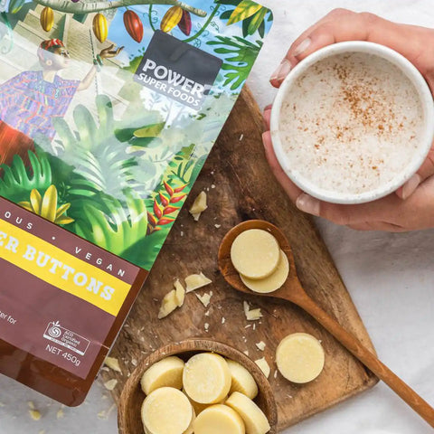 Power Super Foods Cacao Butter Buttons lifestyle shot of the pouch beside a bowl of white buttons and a hand holding a mug, styled lifestyle photo