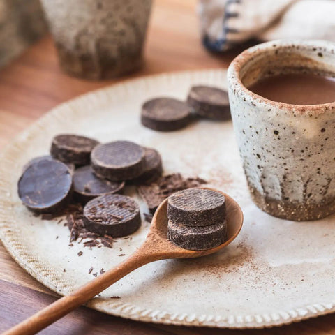 Power Super Foods Ceremonial Cacao Paste Raw Buttons lifestyle shot of dark discs on a plate with a wooden spoon and a cup of hot chocolate