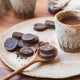 Power Super Foods Ceremonial Cacao Paste Raw Buttons lifestyle shot of dark discs on a plate with a wooden spoon and a cup of hot chocolate
