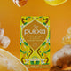 Pukka lemon, ginger, and manuka honey tea box on an orange background with lemons and ginger.