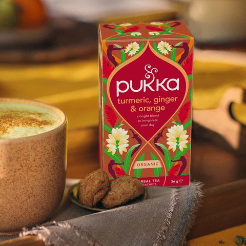 Pukka turmeric, ginger & orange tea box with a cup of tea and cookies on a wooden table.