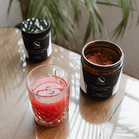 Two containers of Qsilica Vegan Collagen on a wooden surface with a glass of red liquid.