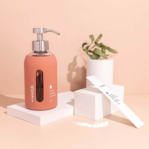 Rose-coloured hand soap dispenser with pump on a white stand against a peach background.