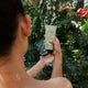Person holding a skincare product under a shower with lush greenery in the background.
