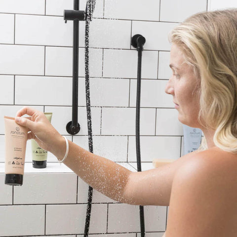 Woman in a shower holding a tube of skincare product with a tiled bathroom background.