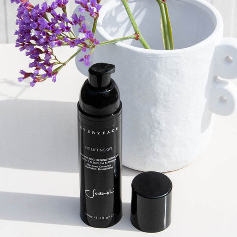 Eye lifting gel with the cap off, positioned in front of a white pot with purple flowers.