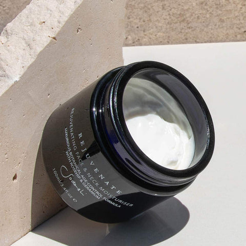 Black jar of rejuvenate face and neck moisturiser on a light stone surface.