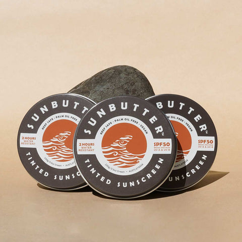 Three tins of SunButter tinted sunscreen on a beige background with a rock.