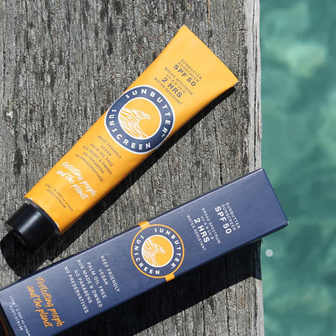 SunButter Oceans Sunscreen 120g yellow tube next to blue packaging on a wooden surface next to the sea.