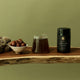 Jar of coffee, glass of coffee, and bowl of figs on a wooden surface with a green background