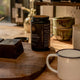 SuperFeast Mason's Mushrooms Blend lifestyle shot of the black jar on a wooden board beside dark chocolate, mugs and a table lamp