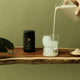 Person pouring a liquid into a glass with ice cubes on a wooden surface, with a green background.