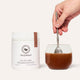 Sleep Ritual container next to a glass of the chocolate oral powder being mixed in with a spoon. Set against white background. 